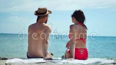 穿着泳装、坐在沙滩上、在沙滩上放松、欣赏阳光明媚的海景的微笑情侣